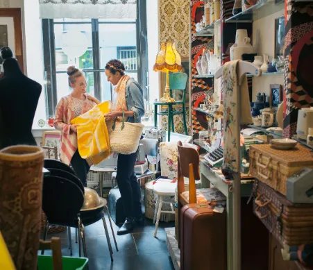 Two women stand with a yellow table cloth in a secondhand store,